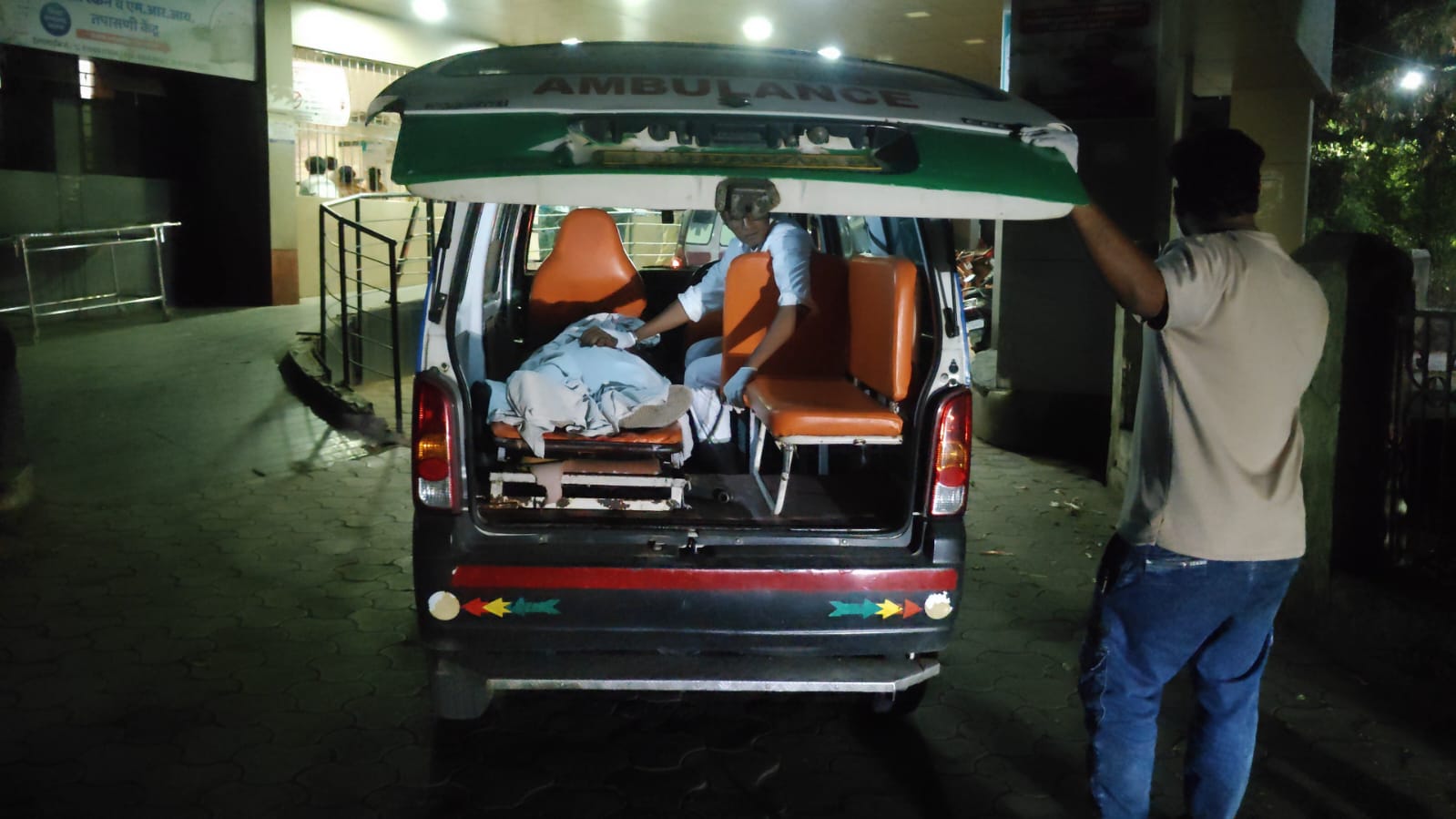 Ambulance with open back doors; a patient on a stretcher being attended by a caregiver inside, at night outside a hospital entrance.