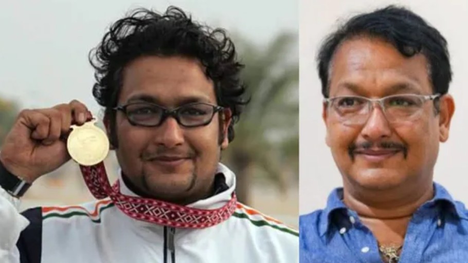 Two men standing side by side; the left man proudly shows a gold medal toward the camera, wearing a patterned scarf and glasses, while the right man smiles in a blue shirt and glasses.