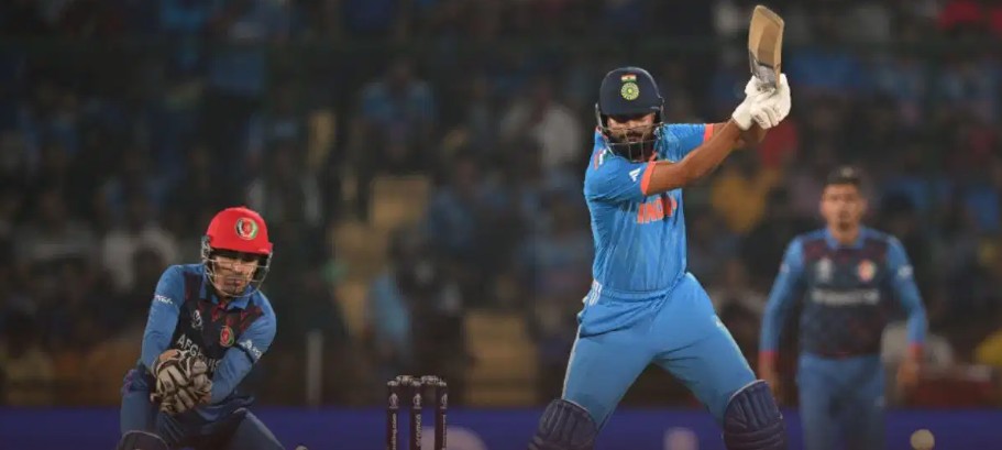 Indian batsman in blue on strike, driving the ball as the wicketkeeper in a red helmet crouches behind the stumps behind him.