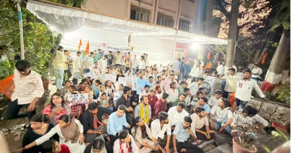 Screenshot_216 - NewsNation Online Large crowd seated on the ground at an outdoor rally under white tents, holding signs and Indian flags in the background.