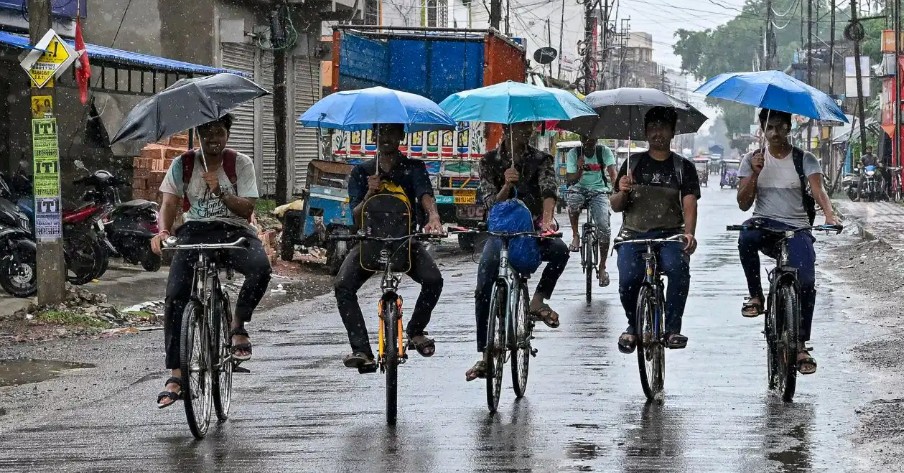 Five people ride bicycles down a wet, rain-soaked urban street, each holding a blue umbrella as they pedal together.