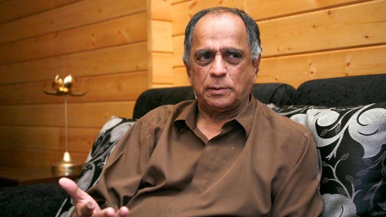Man in a brown shirt seated on a patterned sofa in a wood-paneled room, gesturing with his hand while speaking.