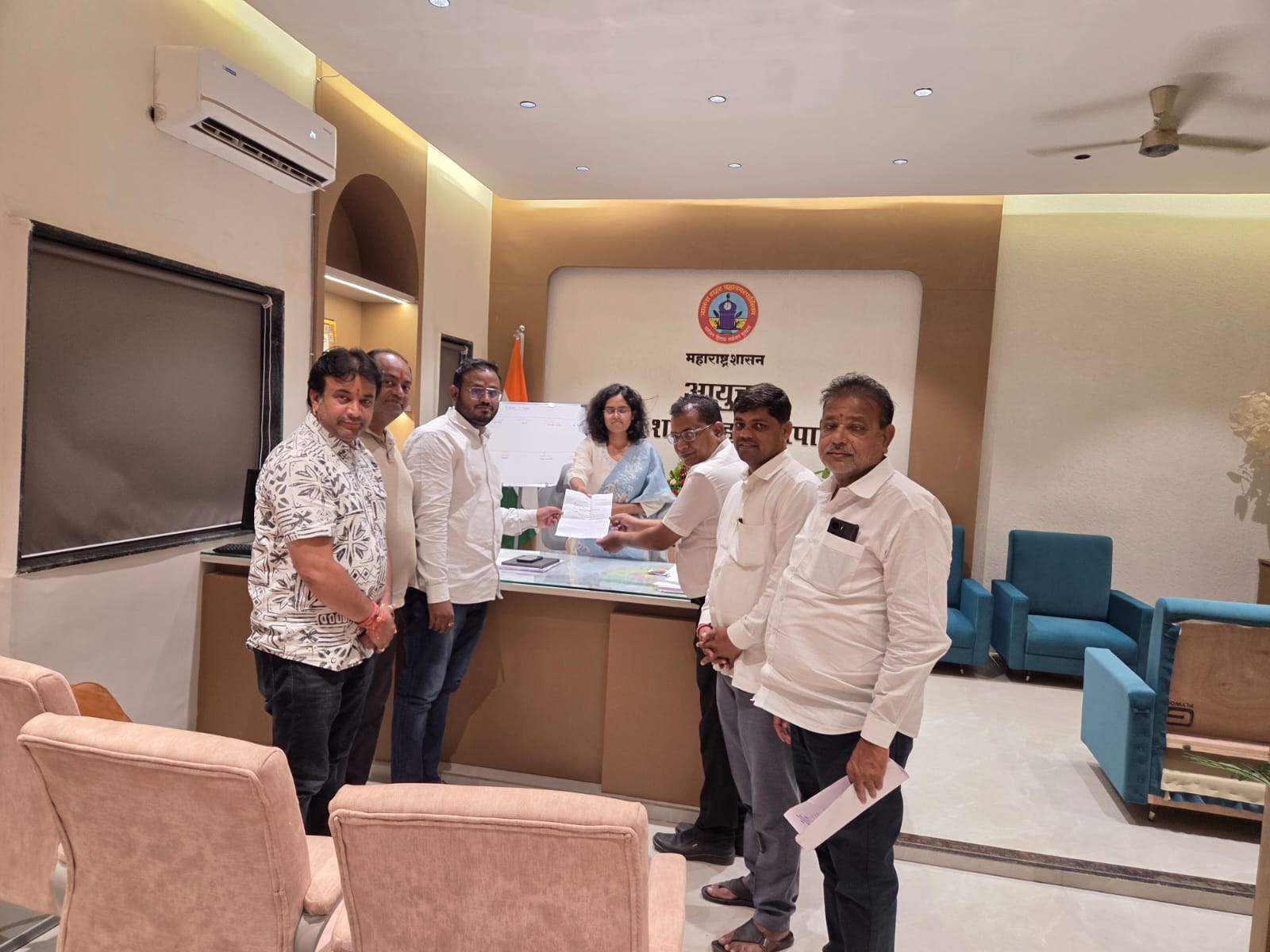 WhatsApp Image 2026-07-18 at 62226 PM - NewsNation Online Group of people standing at a reception desk exchanging a document during a formal office event, with a government emblem on the wall behind them.