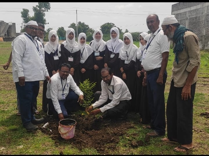 Group of students in white hijabs and mentors planting a sapling in a dirt hole on a grassy field during a tree-planting event. A pink bucket sits nearby.