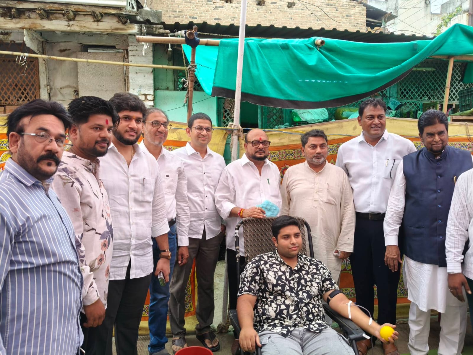 Group of men posing outdoors at a community event; a man in a floral shirt sits in a chair with a medical tube in his arm (blood donation) while others stand behind.