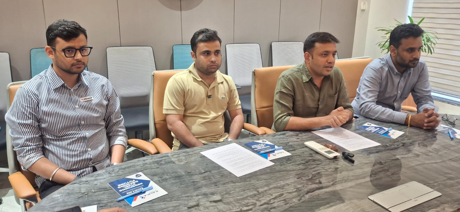 Four men sit at a marble conference table with documents and blue brochures during a meeting in a boardroom.
