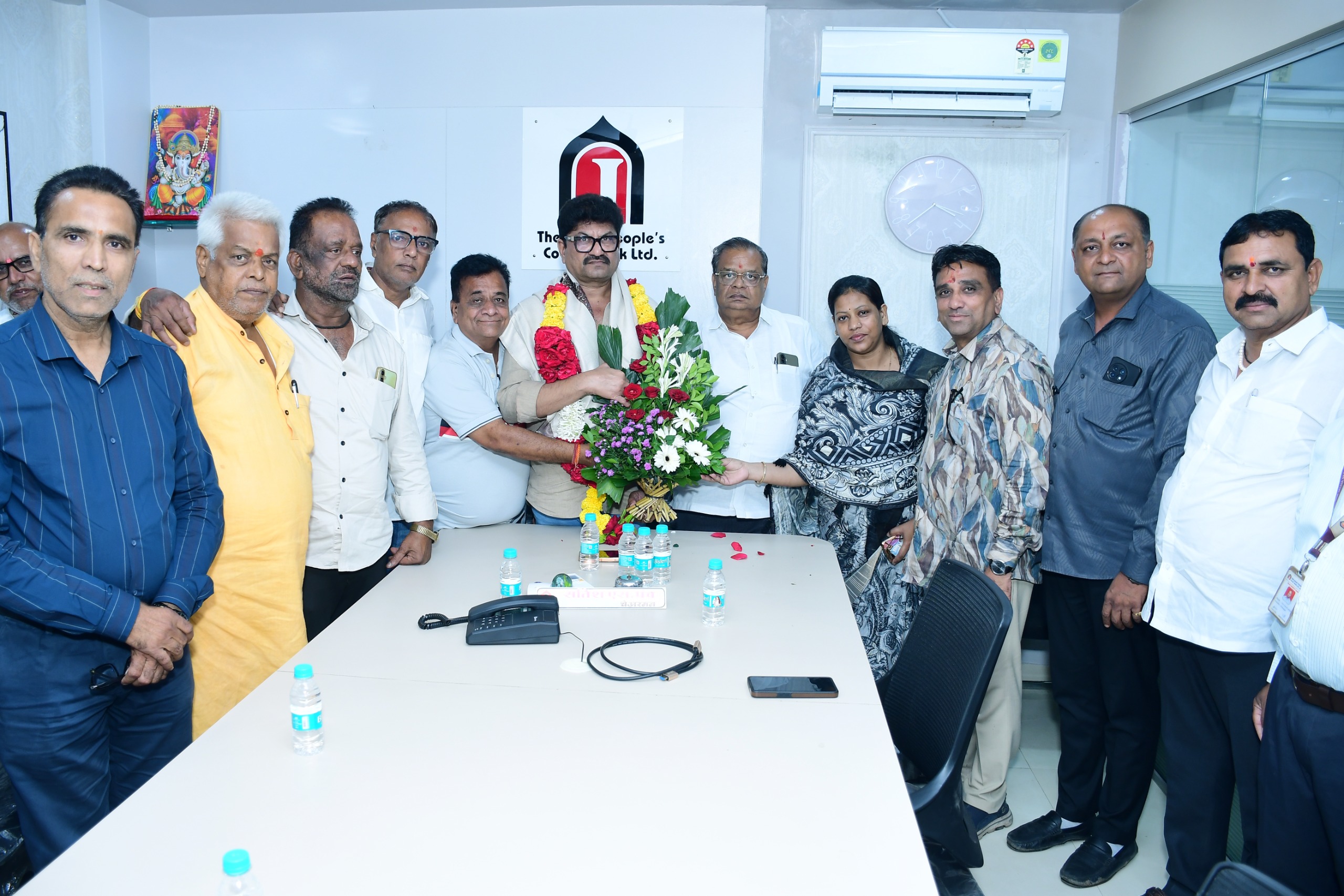 Group of colleagues posing in a conference room; a central man wears flower garlands as others present a bouquet, celebrating an event.