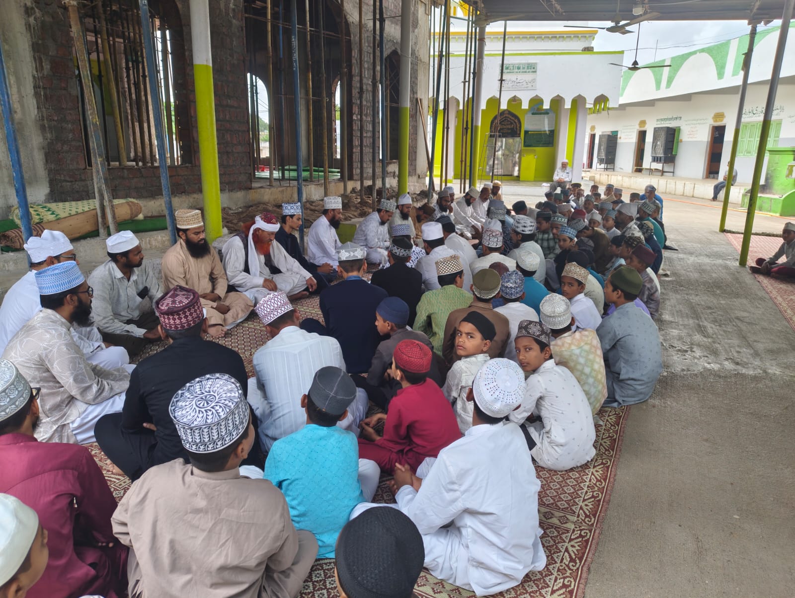 A large group of Muslim men and boys sit cross‑legged in a circle on patterned carpets inside a mosque courtyard, many wearing traditional caps and white robes. construction scaffolding and green pillars are visible in the background.