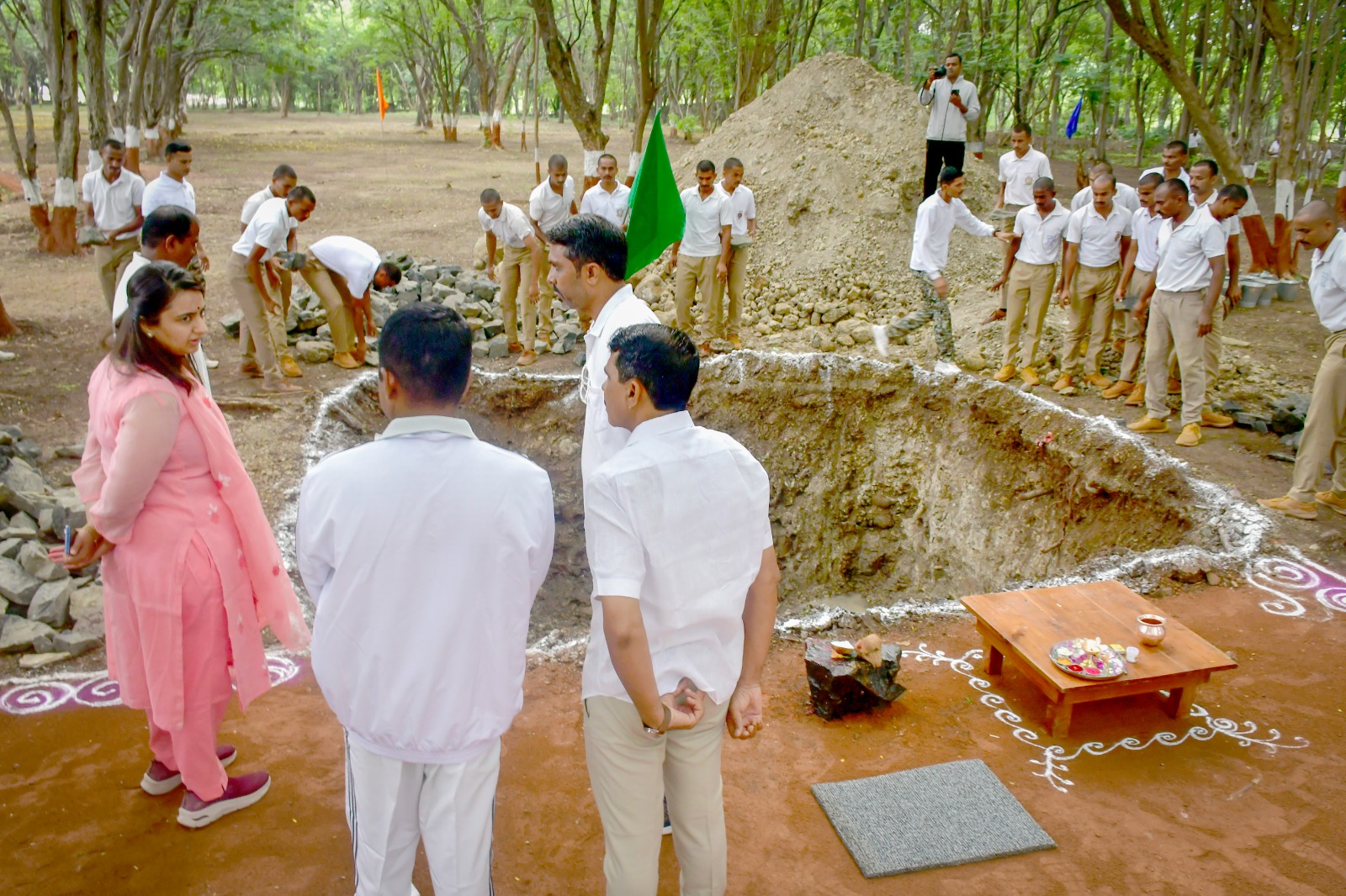 Group of students in white shirts and beige pants gathered around a large dug pit for a groundbreaking ceremony in a wooded area.