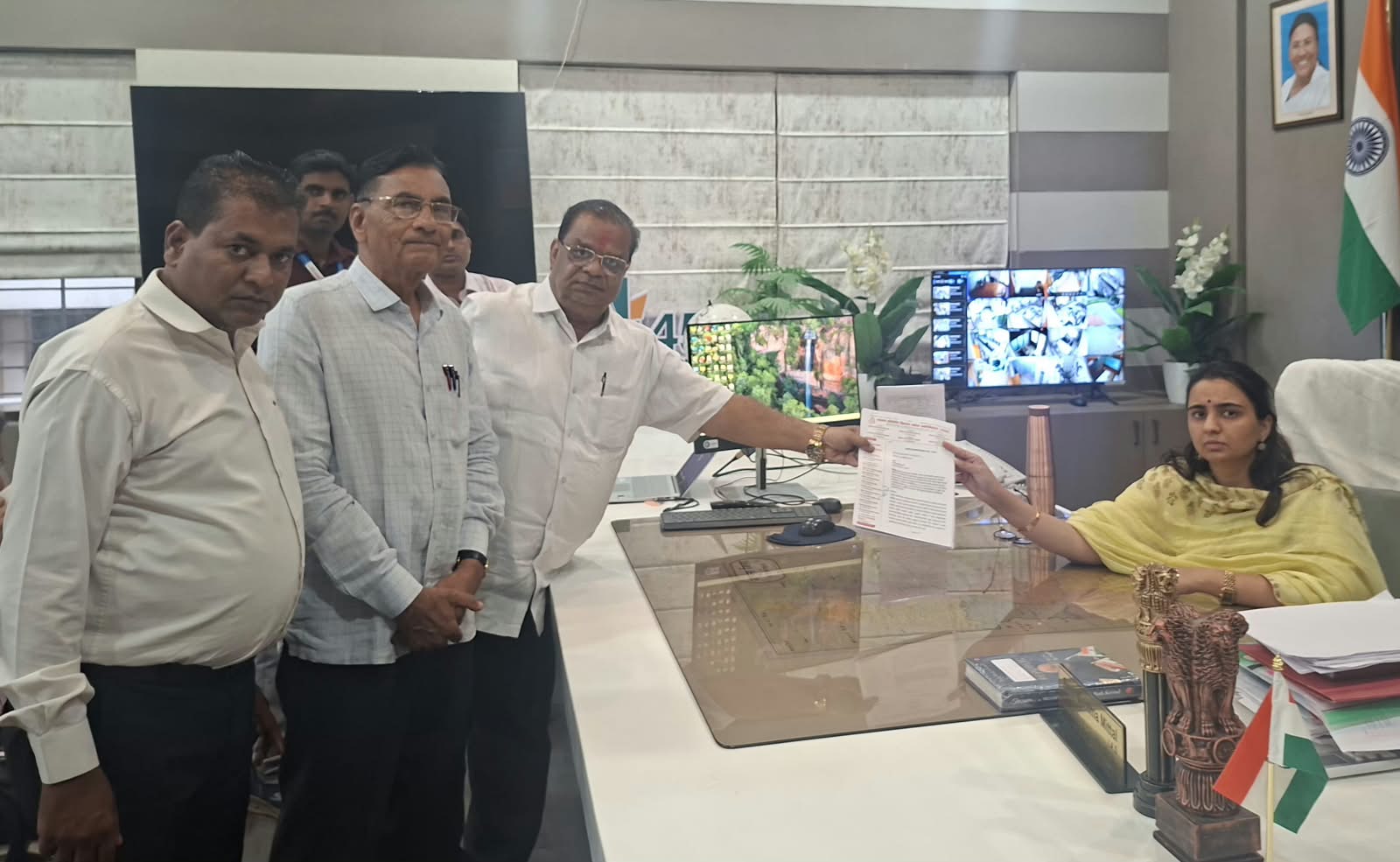 Group of men standing at a reception desk as a woman seated hands over a document in an office setting with Indian flag.