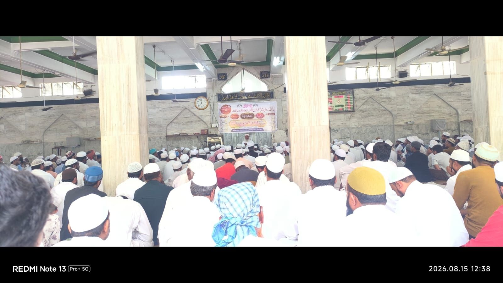 Large crowd of Muslim men seated on the floor inside a mosque, facing a preacher on a stage with a banner.