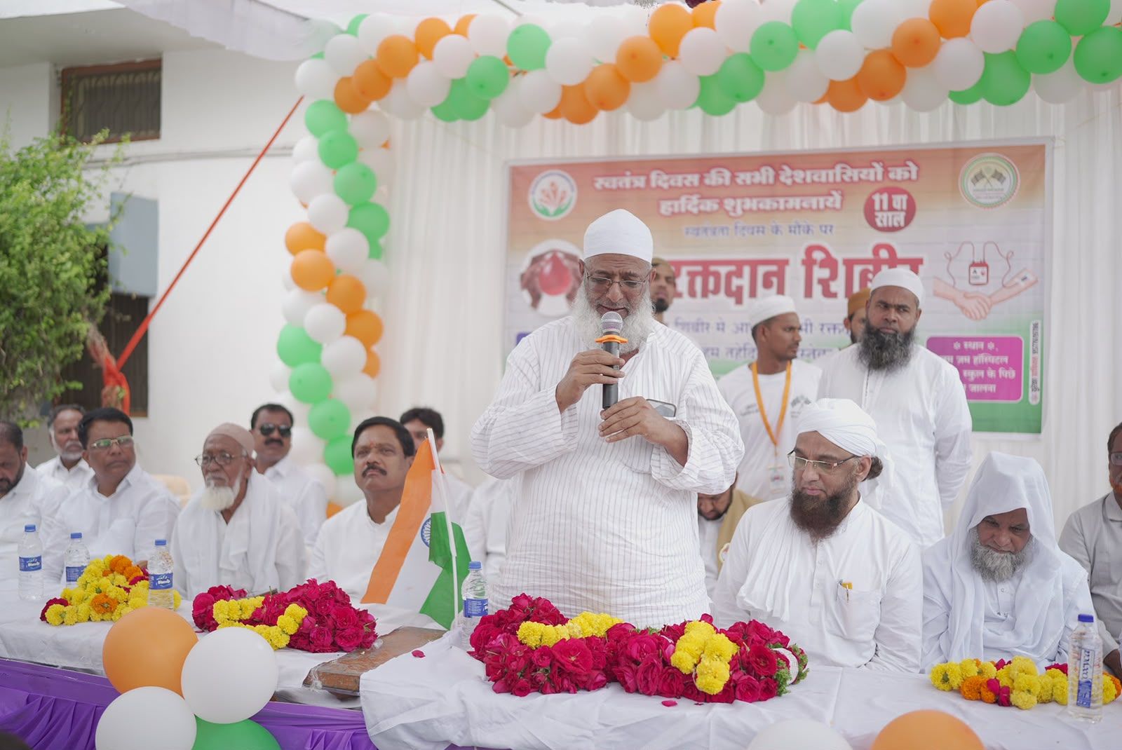 Man in white robe speaking into a microphone at a decorated outdoor ceremony, with the Indian flag on the table and flower garlands in front.