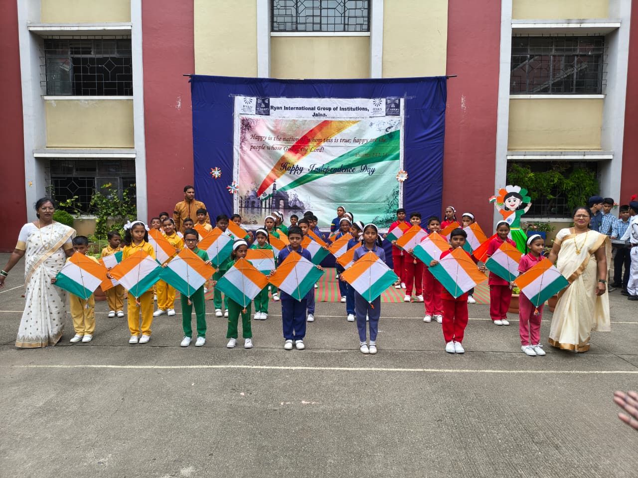 Group of schoolchildren in colorful uniforms holding diamond-shaped Indian flags during an Independence Day celebration in a school courtyard, with teachers nearby.