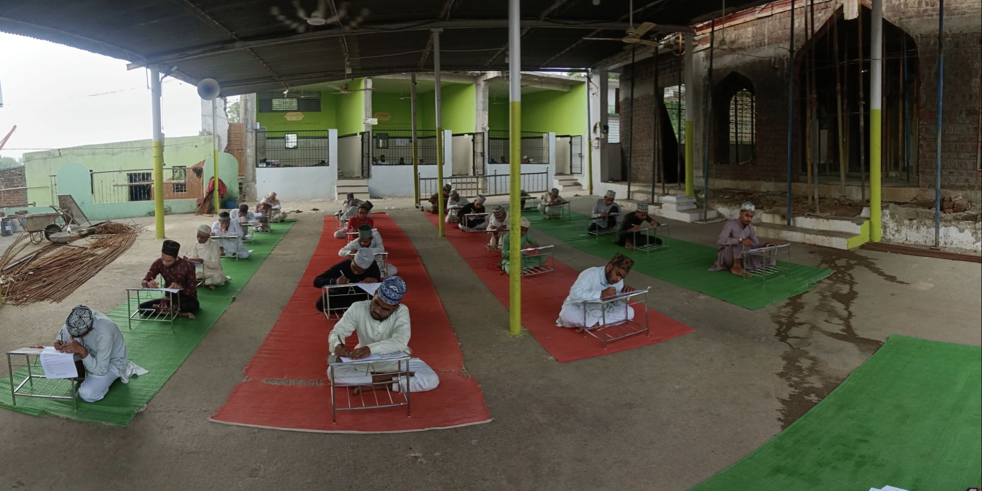 Men in traditional clothing sit on red and green mats, writing at individual desks in a covered open-air hall, likely an exam setting.