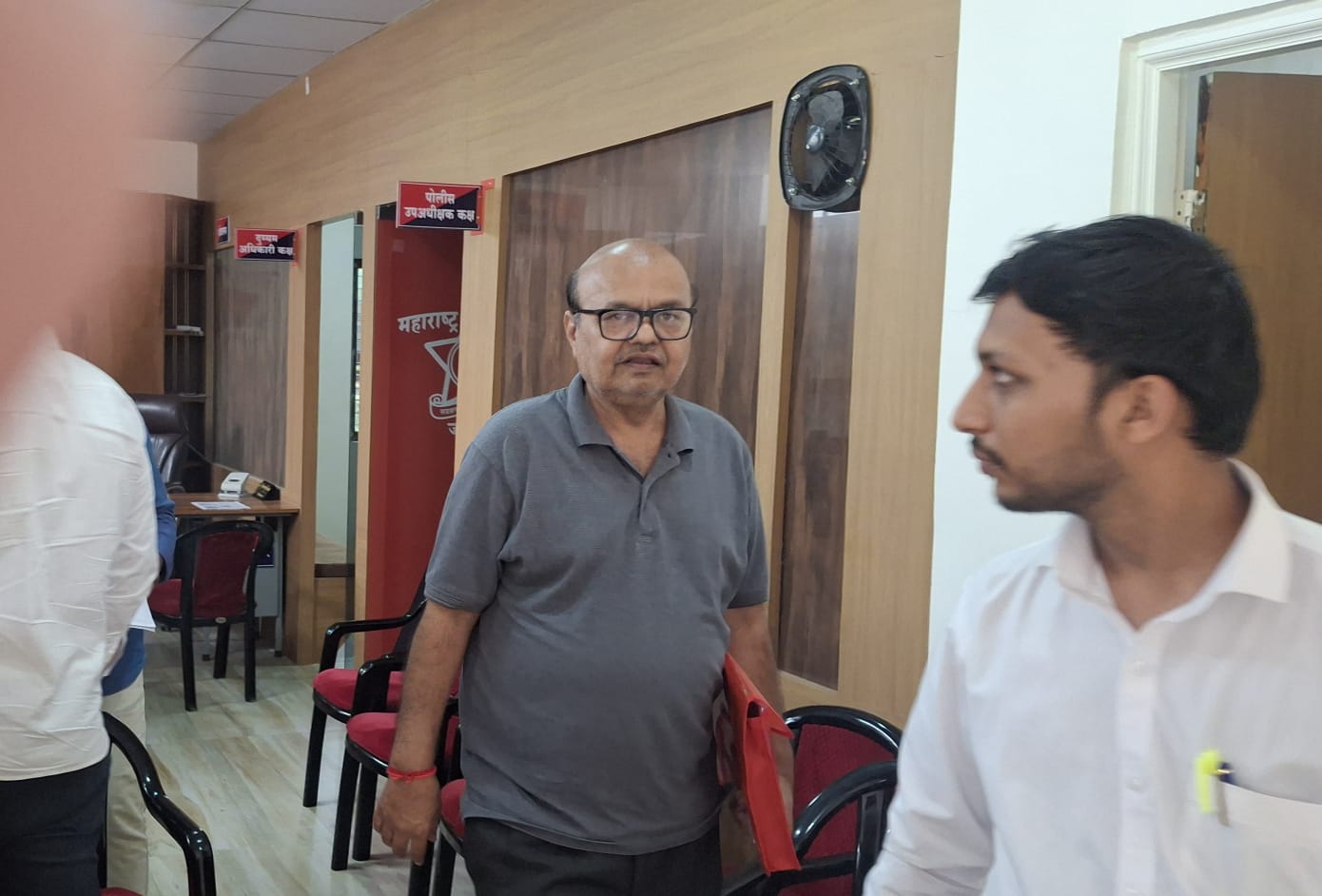 Office lobby scene with two men: one in a gray polo holding a red folder, facing the camera, and another in a white shirt standing to the right; red chairs and wooden panel walls in the background.