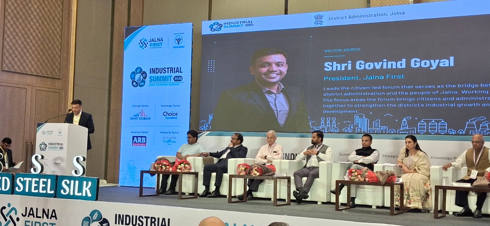 Speaker at a podium addressing a panel on stage during an Industrial Summit, with banners and a large screen in the background.