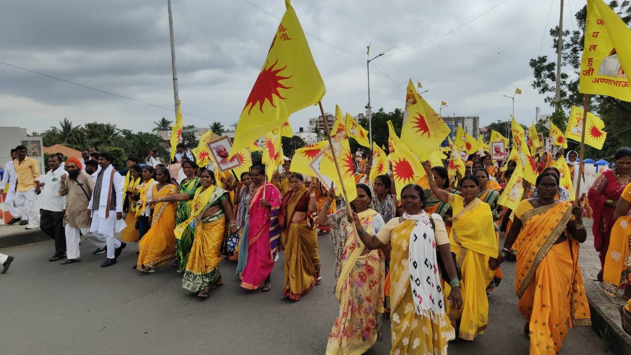 Group of people marching in a street protest carrying yellow flags with a red sun emblem, many wearing colorful sarees.
