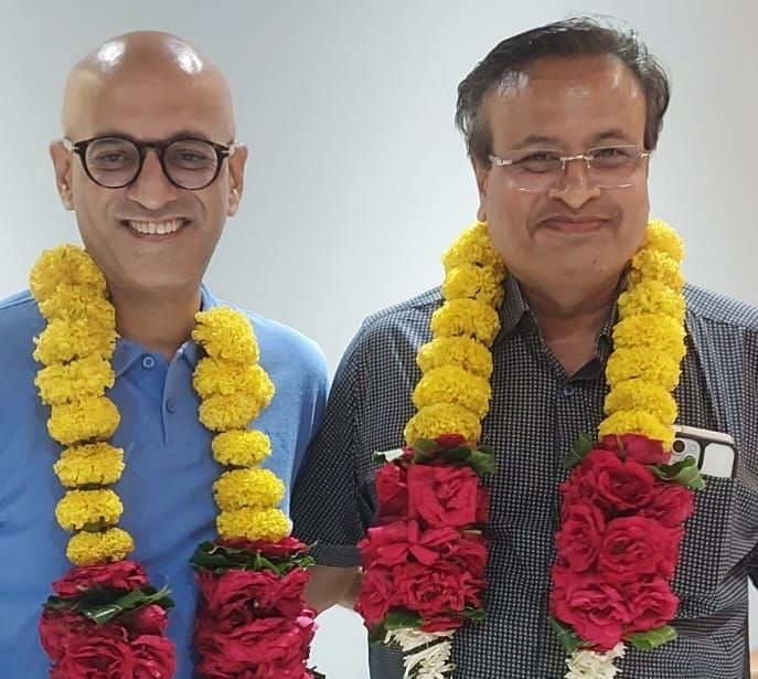 Two men wearing yellow marigold and red rose garlands, smiling at the camera indoors.