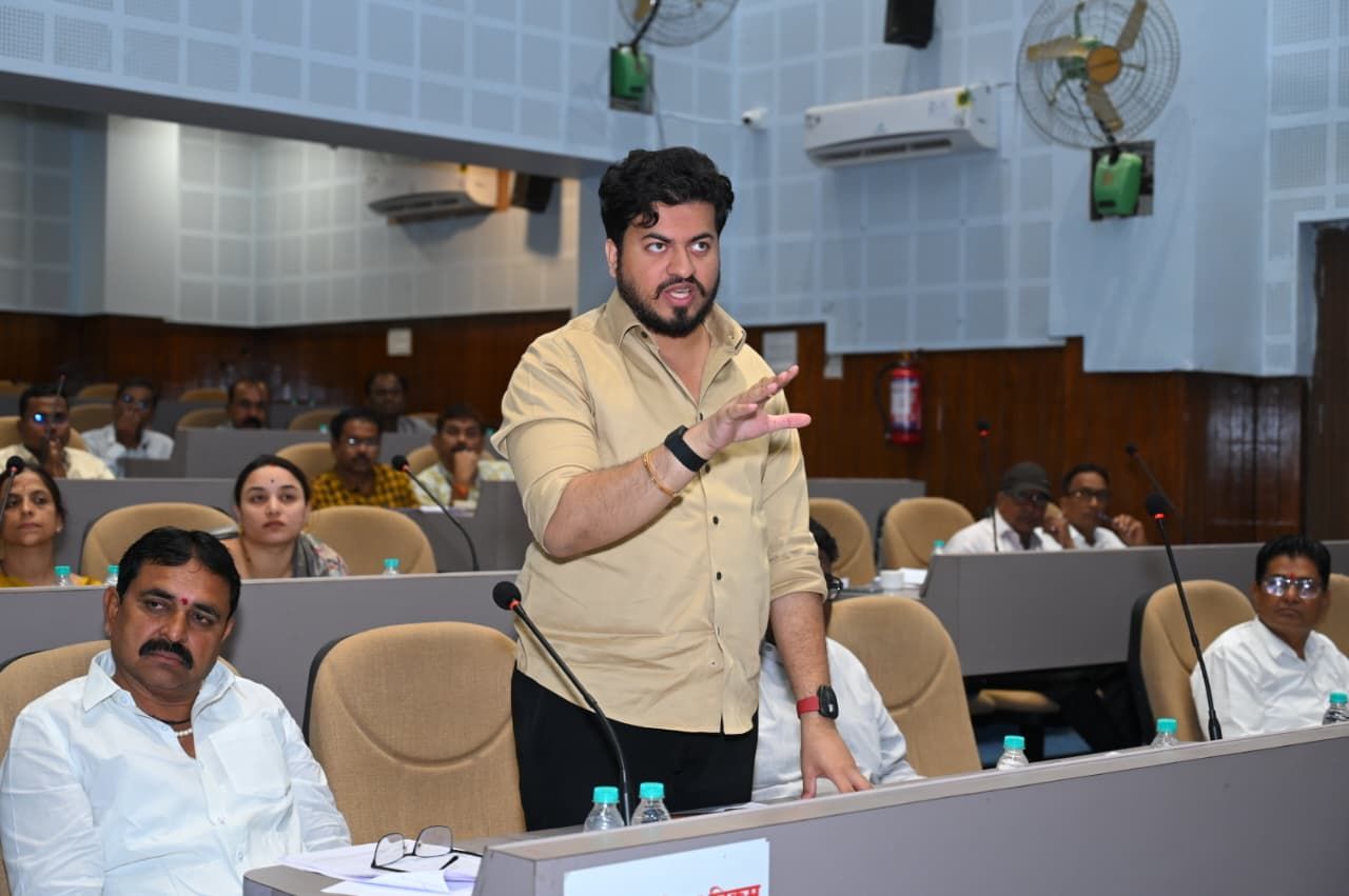 Man in a beige shirt stands and speaks at a conference podium while a seated audience listens in a lecture hall.