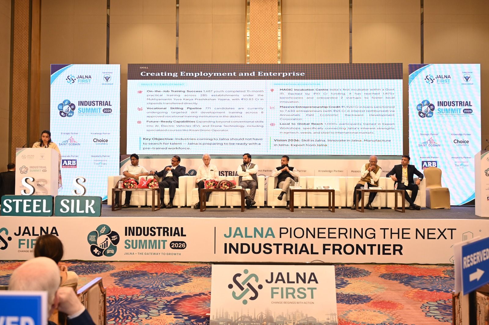 Panelists seated on stage during the Industrial Summit 2026, with a presenter at a podium on the left and a large screen behind them showing event details.