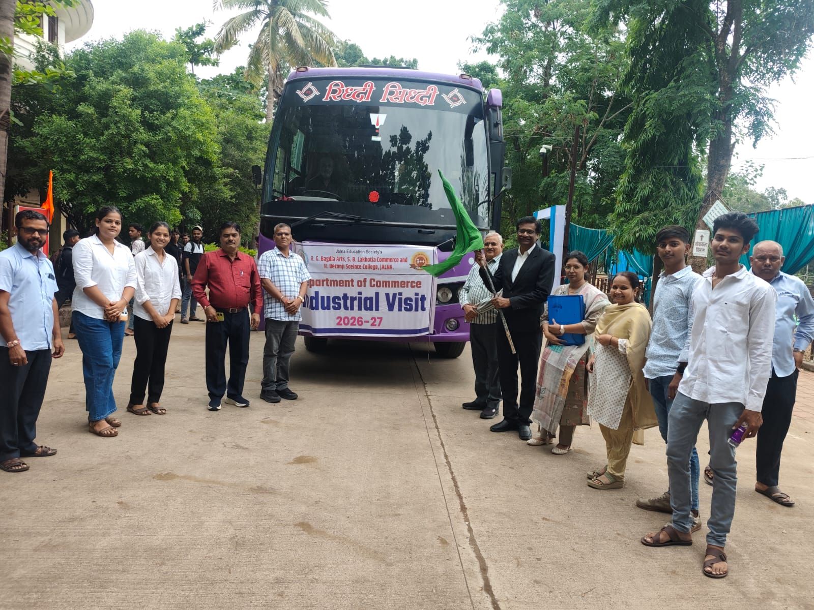 Group of students and staff standing beside a purple bus with an 'Industrial Visit 2026-27' banner; campus trees in background.