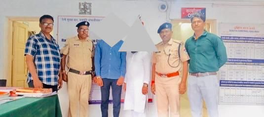 Six people, including police officers, pose for a photo indoors; two civilians in the center wear blue and white, their faces obscured by a gray triangle; a green table and information boards are in the background.