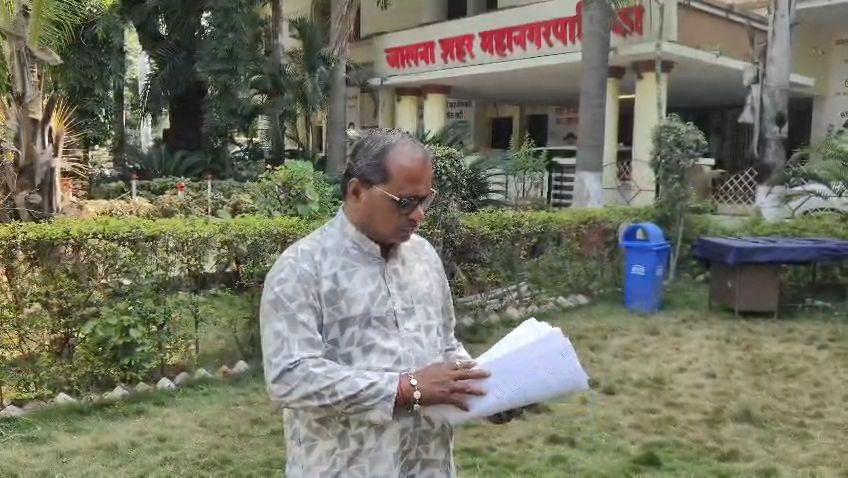 Man in a patterned kurta and sunglasses stands in a garden, reading a stack of papers outside a building.