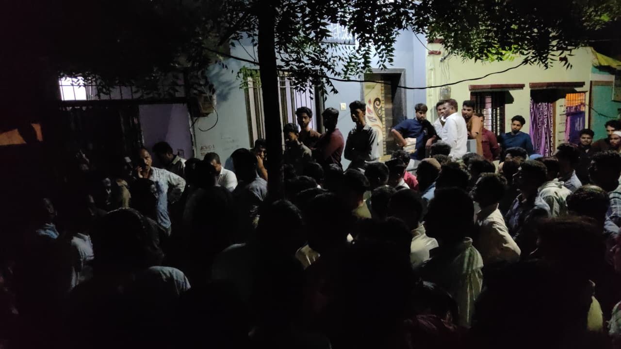img-20260919-wa00132316762205017669216 - NewsNation Online Night-time street gathering: a dense crowd of people standing and talking outside a row of houses under tree branches.