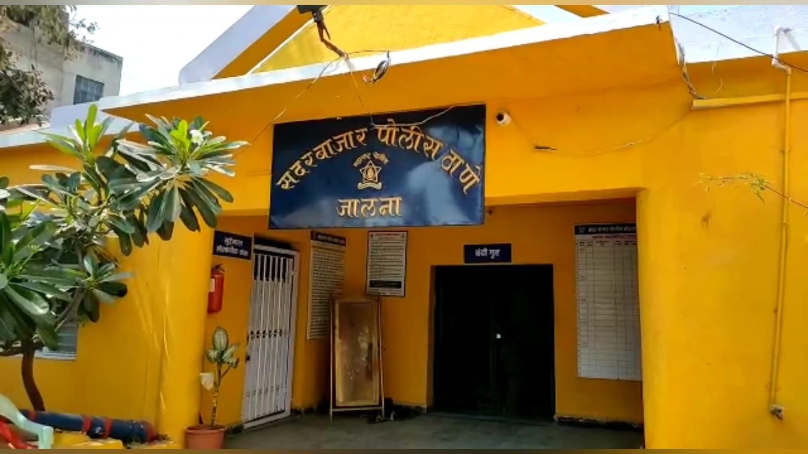 img-20260920-wa00151268738985380664297 - NewsNation Online Entrance to a bright yellow police station with a blue signboard displaying Hindi text and an emblem above the doorway. A metal gate and a bulletin board flank the entrance, with potted plants nearby.