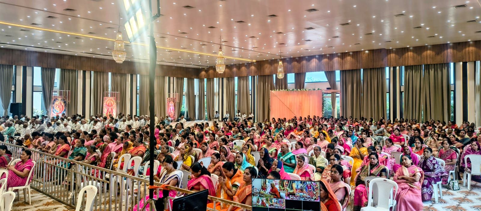 Large hall filled with people seated for a cultural or religious gathering; many women wear colorful sarees in pink, orange, and green.