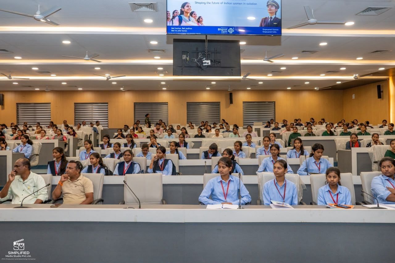 Large lecture hall filled with students in light blue uniforms seated at desks with microphones, facing the front where a presentation screen hangs above.
