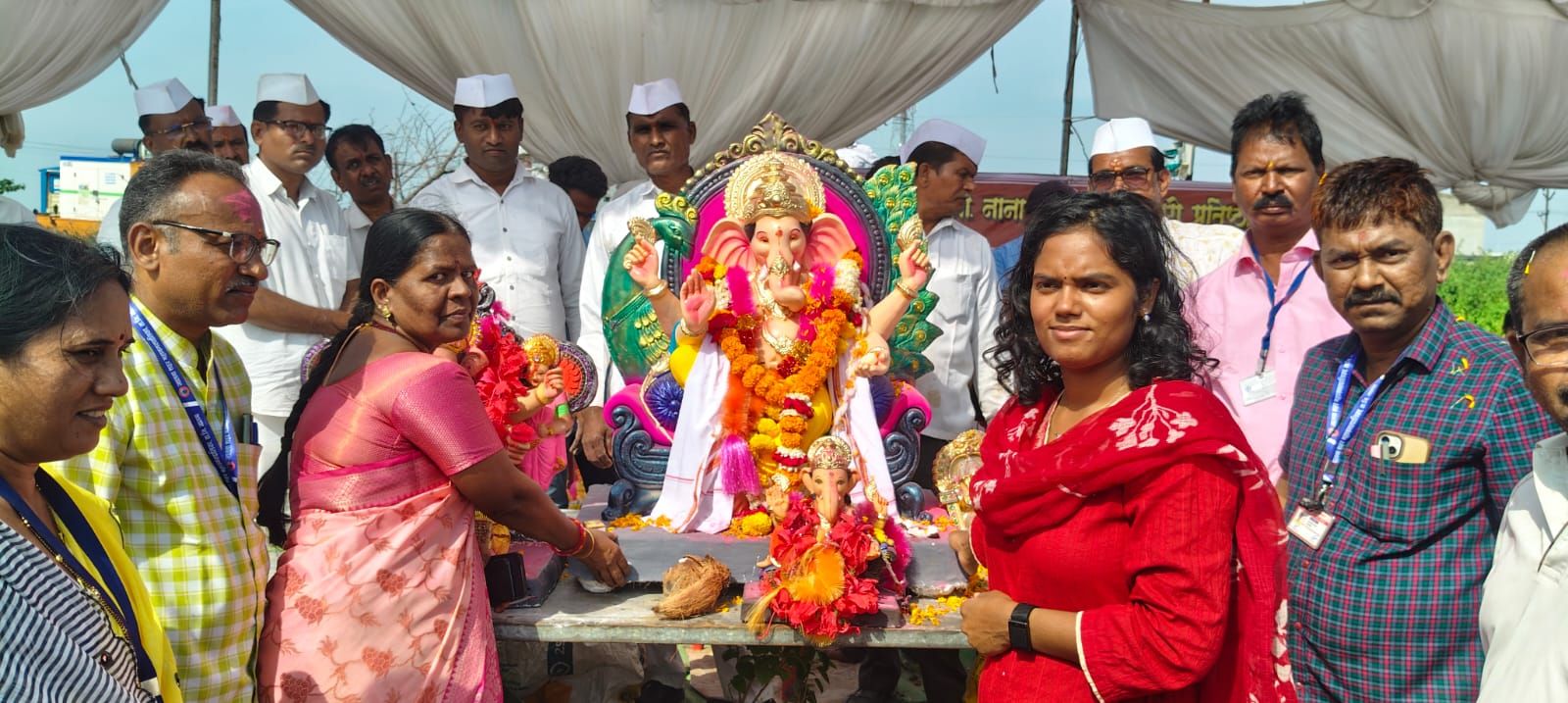 People gathered around a large, garlanded statue of Lord Ganesha with offerings, under a white canopy.