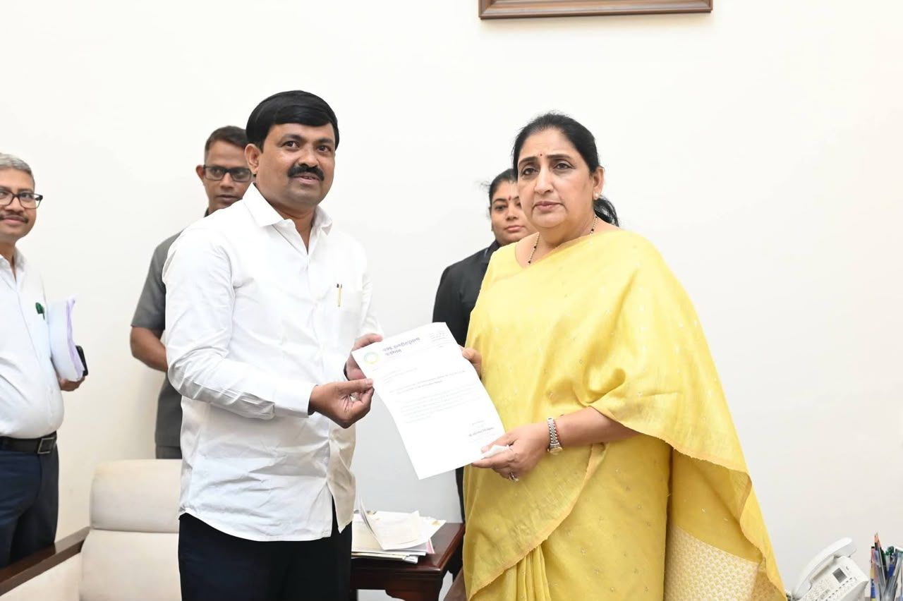 Man in a white shirt hands a document to a woman in a yellow saree in an office, with colleagues in the background.