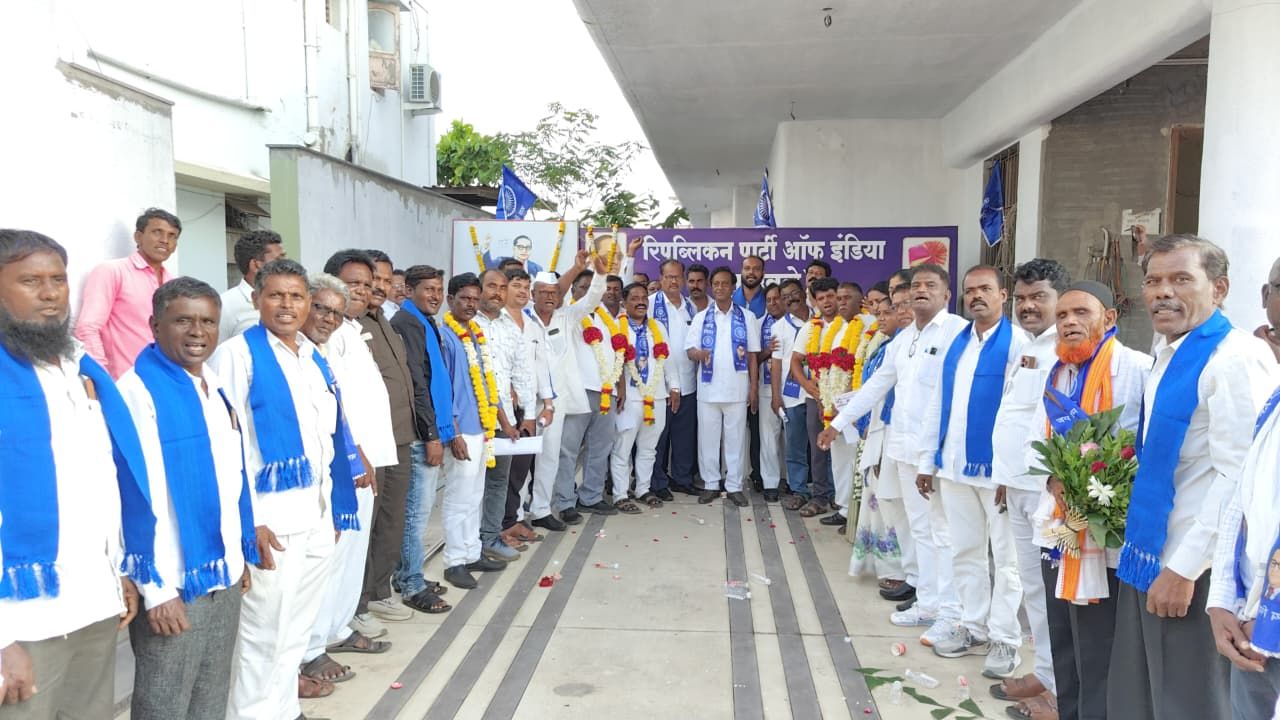 Group of men in white outfits with blue scarves lined up in a corridor, some with flower garlands, posing for a photo at a political event with a banner in the background.