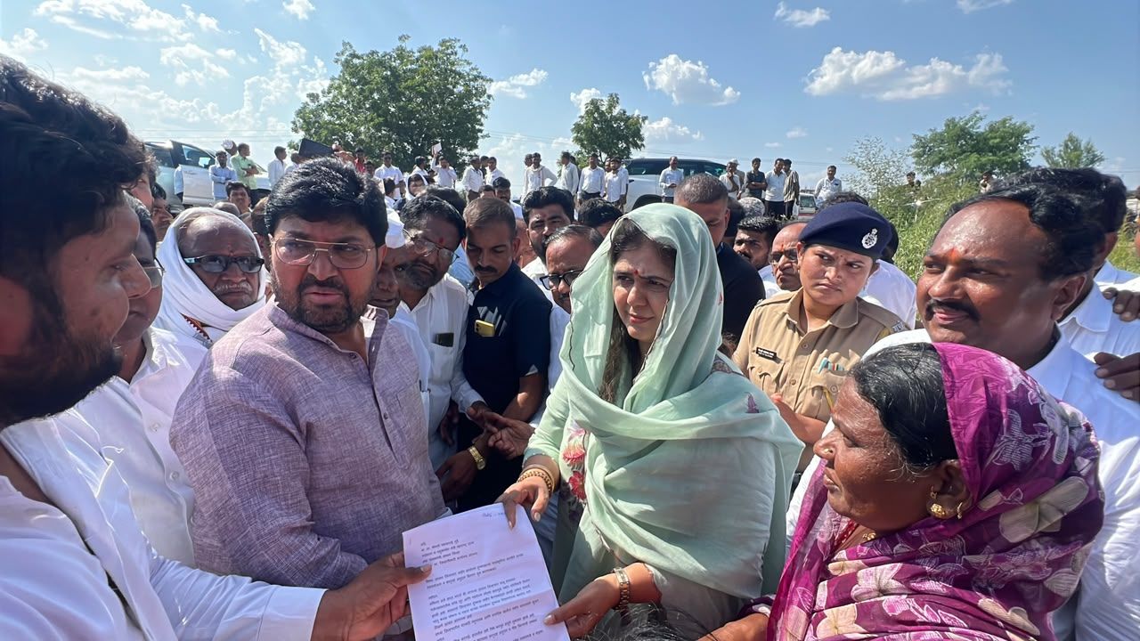 Woman in a light green scarf hands a document to a man as a crowd of officials and villagers gathers outdoors on a sunny day.