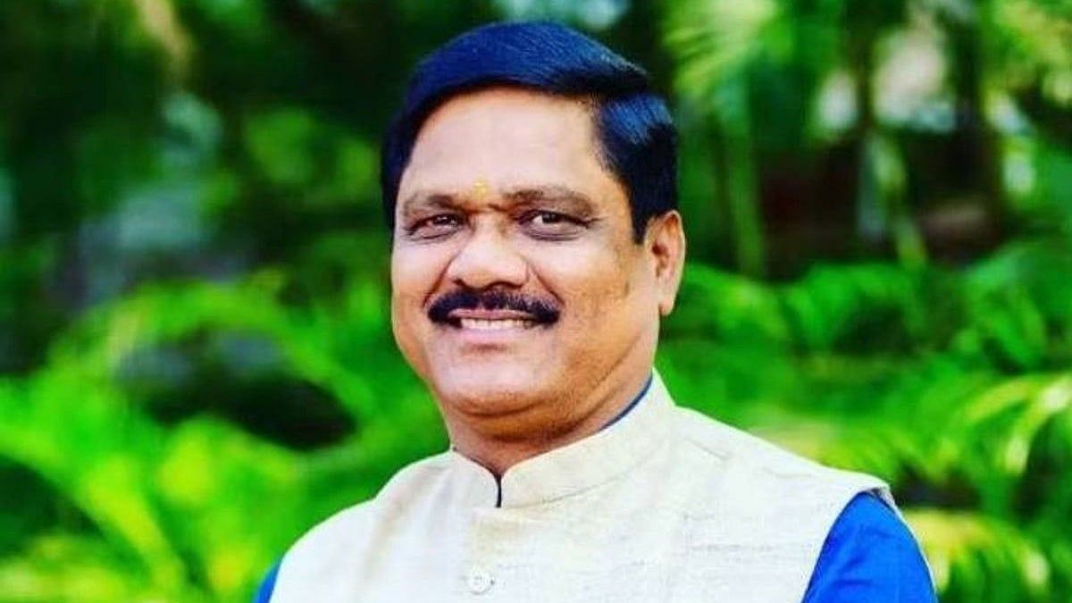 Portrait of a smiling man in a cream Nehru jacket with blue sleeves, outdoors among lush green plants.