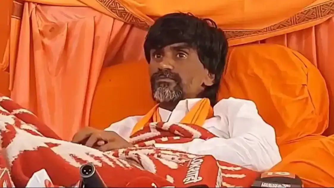 Man with dark hair and gray beard, in a white shirt and orange scarf, reclining on an orange couch under draped fabric, covered by a red-and-white blanket.