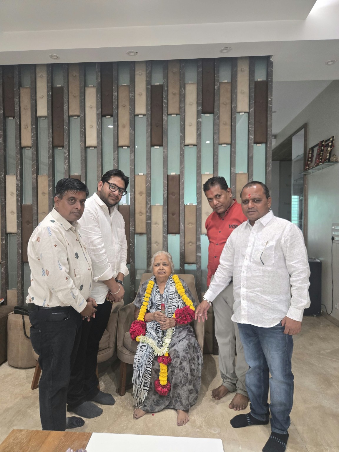 Elderly woman seated with garlands around her neck, flanked by five men in a ceremony-like gathering indoors.
