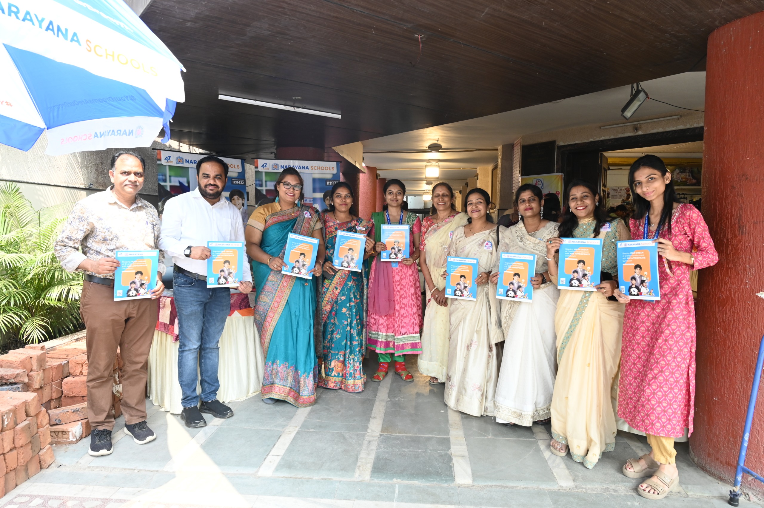 IMG-20261026-WA0017 - NewsNation Online Group of men and women in traditional Indian attire smiling and holding blue-and-orange booklets at an outdoor school event under a canopy, with Narayana Schools banners in the background
