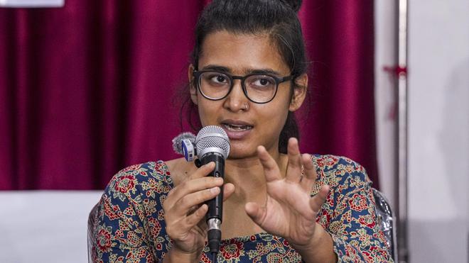 Woman with glasses speaks into a microphone, gesturing with her hand during a talk on stage with burgundy curtains behind her.