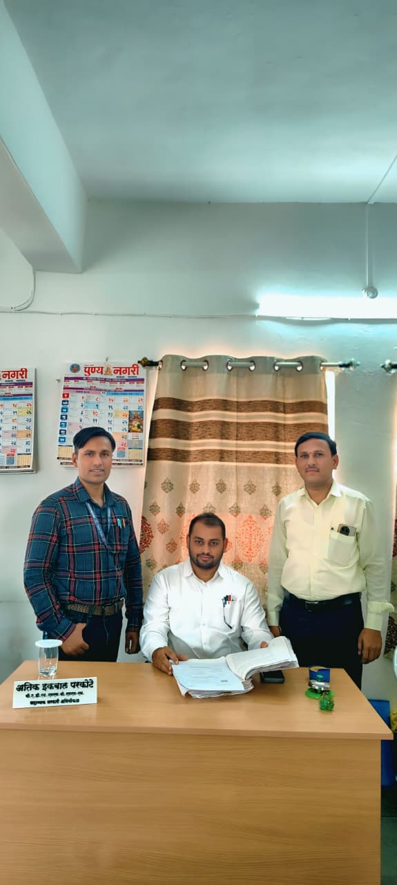कार्यालय में मेज पर दस्तावेजों के साथ बैठे एक व्यक्ति और पास खड़े दो अन्य व्यक्ति